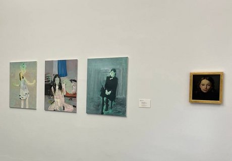 Auf der bunten Fotografie sieht man einen Ausstellungsraum mit weißen Wänden. An einer Wand hängen verschiedene Portraits von jungen Frauen. Hinten sieht man einen Flur. An der weißen Wand dort hängt eine Fotografie.