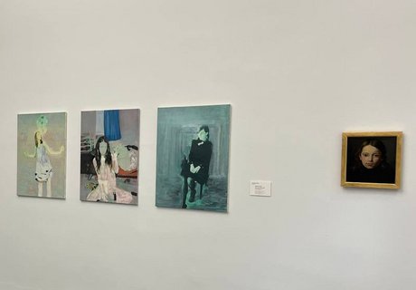 Eine bunte Fotografie. Darauf ist ein Ausstellungsraum mit weißen Wänden zu sehen. An einer der Wände hängen verschiedene Potraits von jungen Frauen. Im Hintergrund ist der Teil eines Flures zu sehen. Dort hängt an der weißen Wand eine Fotografie.