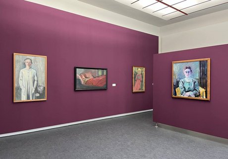 Eine bunte Fotografie. Darauf sind Teile eines Ausstellungsraums des Museumsberg Flensburgs zu sehen. Die Wände sind in einem Fuchsia-Ton gestrichen. An der einen Wand hängen drei Gemälde der Künstlerin Käte Lassen. Auf den drei Gemälden sind unterschiedliche Frauen dargestellt. Auf der Wand rechts befindet sich ein weiteres Portrait von einem anderen Künstler. 