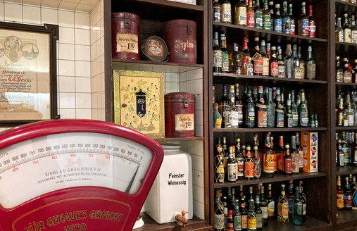 Auf dem Bild ist ein Kolonialwarenladen zu sehen. Ganz links steht eine rote Waage auf dem Tresen, dahinter ist ein Regal mit unzähligen Rumflaschen sowie Tee- und Kaffeedosen.