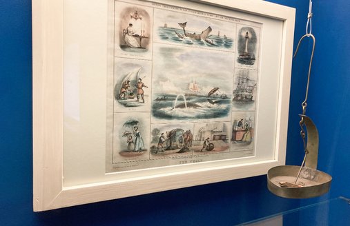In einer Vitrine mit blauer Rückwand hängt eine Zeichnung mit mehreren Bildern über das Thema Walfang. Rechts daneben hängt eine alte Funzel, die mit Tran betrieben worden ist.
