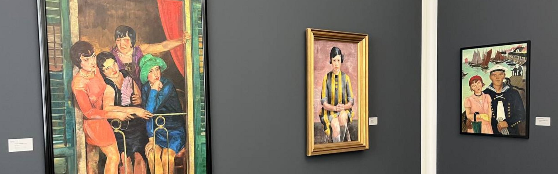 Das Foto zeigt eine Ecke eines Ausstellungsraums. Die Wände sind in einem dunklen Grau gestrichen. An der linken Wand hängen zwei Gemälde. Und an der rechten Wand hängt ein weiteres Gemälde. Das vorderste Gemälde zeigt vier Frauen. Sie tragen Kleider im Stil der 1920er Jahre. Die Frauen stehen auf einem kleinen Balkon und schauen etwas rechts unter sich an. Rechts neben diesem Gemälde ist ein weiteres zu sehen. Es zeigt eine junge Frau, die sitzt und den Betrachter aus dem Bild anschaut. Sie trägt ein gelb-schwarz gestreiftes Kleid. Das nächste Gemälde zeigt eine Frau, die leicht von einem Mann umarmt wird. Die Frau trägt ein rosa Kleid, eine rote Mütze, eine kleine braune Handtasche und einen grünen Regenschirm. Der hängt an ihrem rechten Arm. Der Mann trägt eine Matrosenuniform. Er hat den rechten Arm um die Frau gelegt und hält in der linken Hand weiße Handschuhe. Hinten in diesem Bild sieht man einen Teil eines Hafens mit Holzstegen. Auf dem Wasser sind einige Segelboote und andere Boote zu sehen.