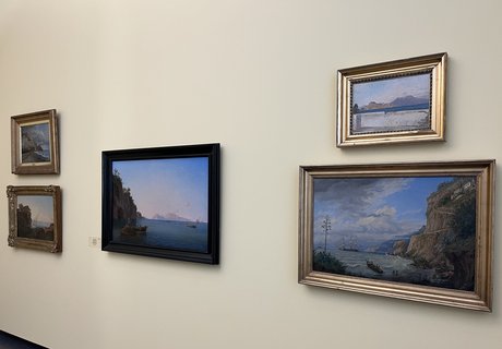 Das Foto zeigt fünf Bilder. Sie hängen an einer hellbeigen Wand. In der Mitte hängt ein Bild in einem schwarzen Rahmen. Links davon hängen zwei Bilder übereinander. Rechts davon hängen auch zwei Bilder übereinander. Auf allen Bildern sind Landschaften mit dem Meer zu sehen.