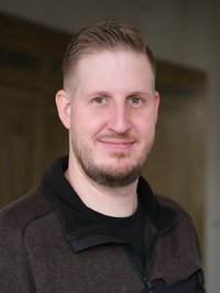 Portrait eines Mannes, der in die Kamera lächelt. Er trägt braune Arbeitskleidung. 