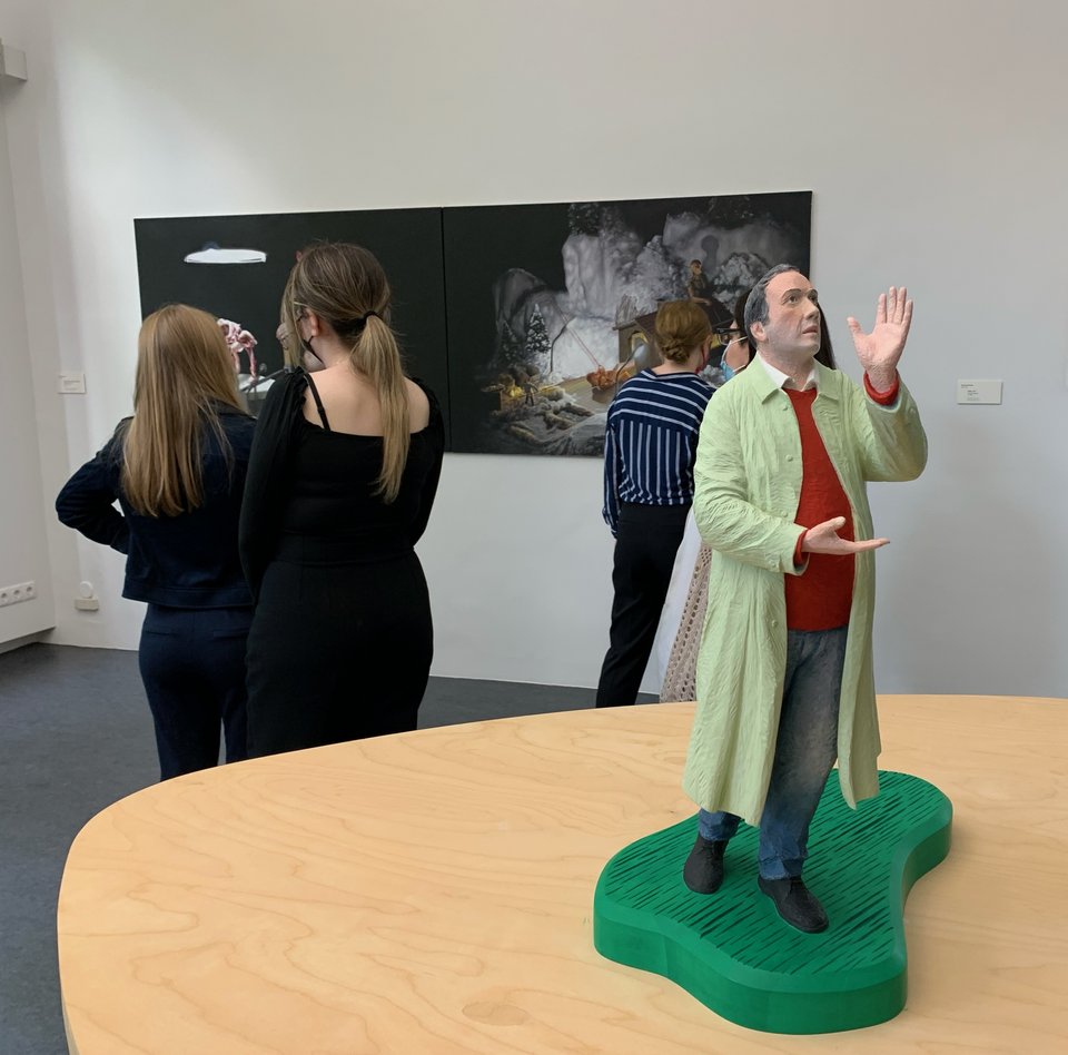 Im Bild befindet man sich in einem der Ausstellungsräume. Im Vordergrund befindet sich die Skulptur eines Mannes im Mantel. Er schaut nach oben. Er gestikuliert mit den Händen. Im Hintergrund befinden sich zwei Gemälde. Vor ihnen stehen drei Besucherinnen, die eins der Gemälde betrachten. 
