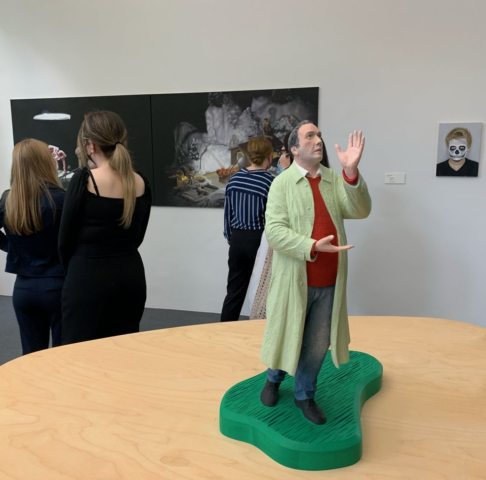 Im Bild befindet man sich in einem der Ausstellungsräume. Im Vordergrund befindet sich die Skulptur eines Mannes im Mantel. Er schaut nach oben. Er gestikuliert mit den Händen. Im Hintergrund befinden sich zwei Gemälde. Vor ihnen stehen drei Besucherinnen, die eins der Gemälde betrachten. 