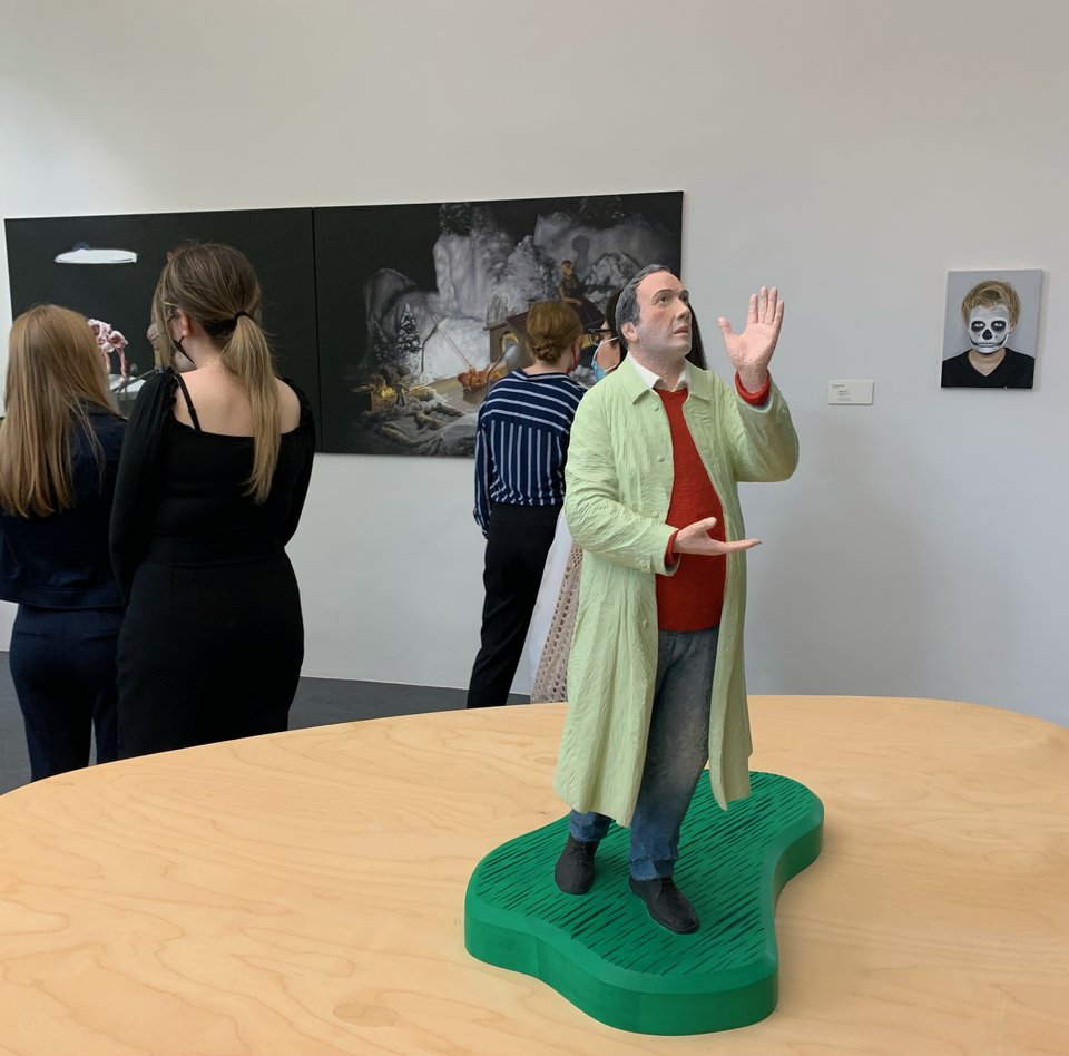 På billedet befinder man sig i et af udstillingslokalerne. I forgrunden står en skulptur af en mand i frakke. Han kigger opad. Han gestikulerer med hænderne. I baggrunden hænger to malerier. Foran dem står tre besøgende, der betragter det ene af malerierne.