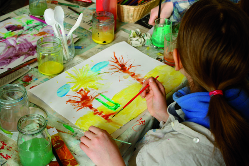 Et barn maler et billede med forskellige farver og pensler. Billedet viser palmer på stranden. På bordet står farvepotter og andet malerudstyr.